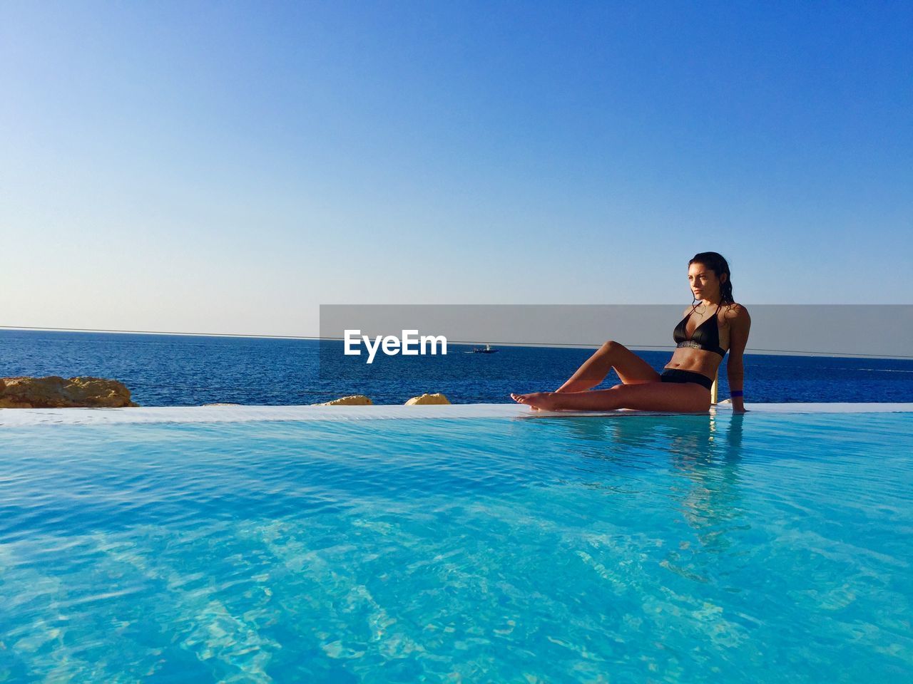 Full length of young woman sitting by swimming pool against sea