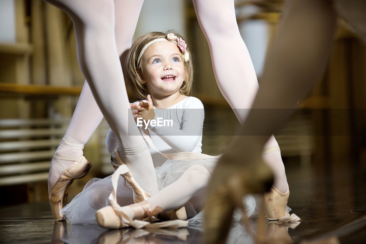 A little ballerina sits on the floor among the legs of adult ballerinas.