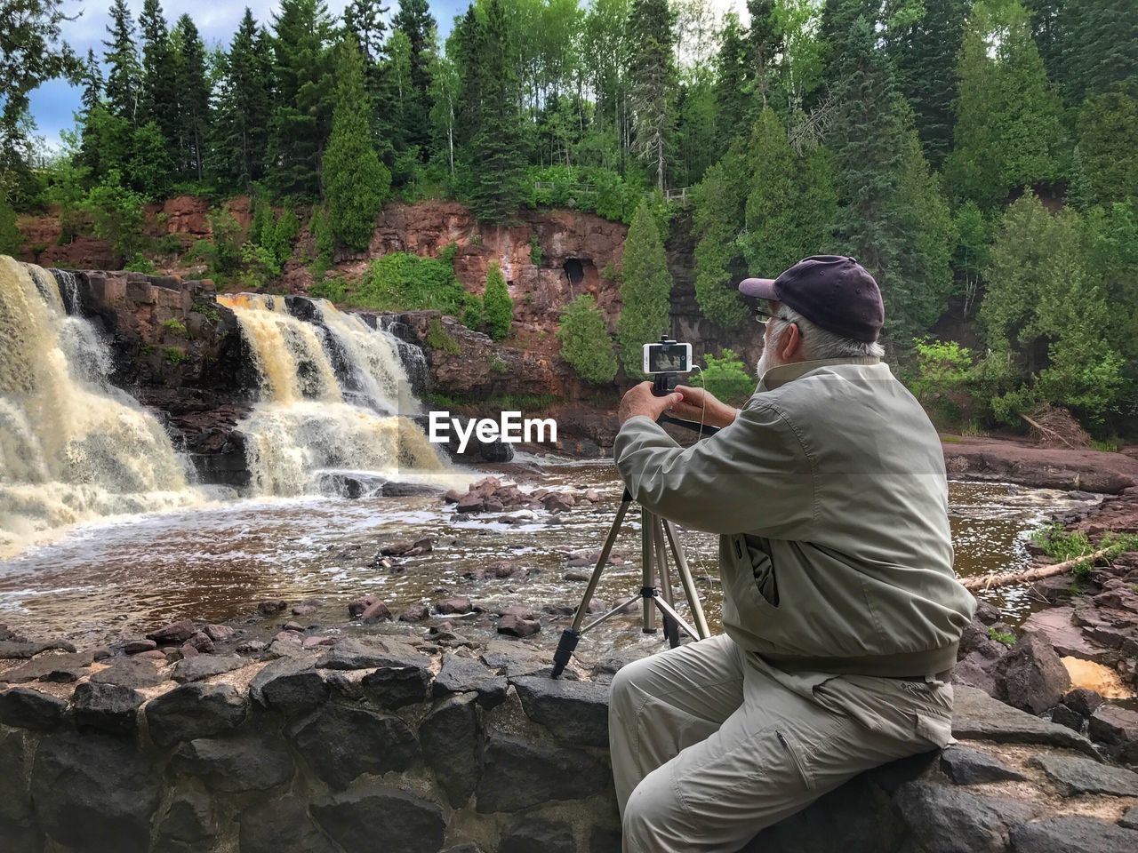 Man photographing waterfall through mobile phone with monopod while sitting at forest