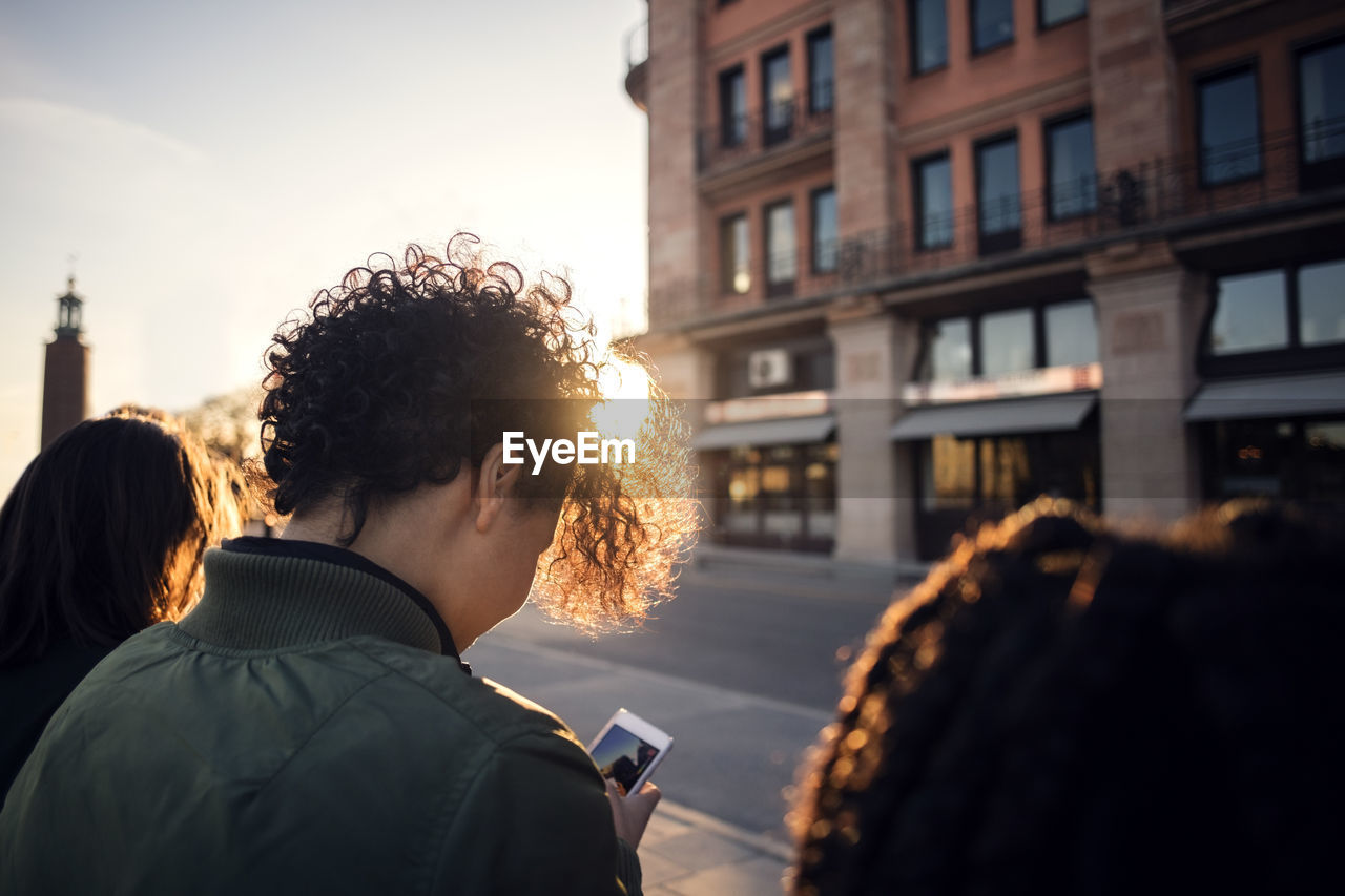 Teenage girl using phone while standing with friends by street in city