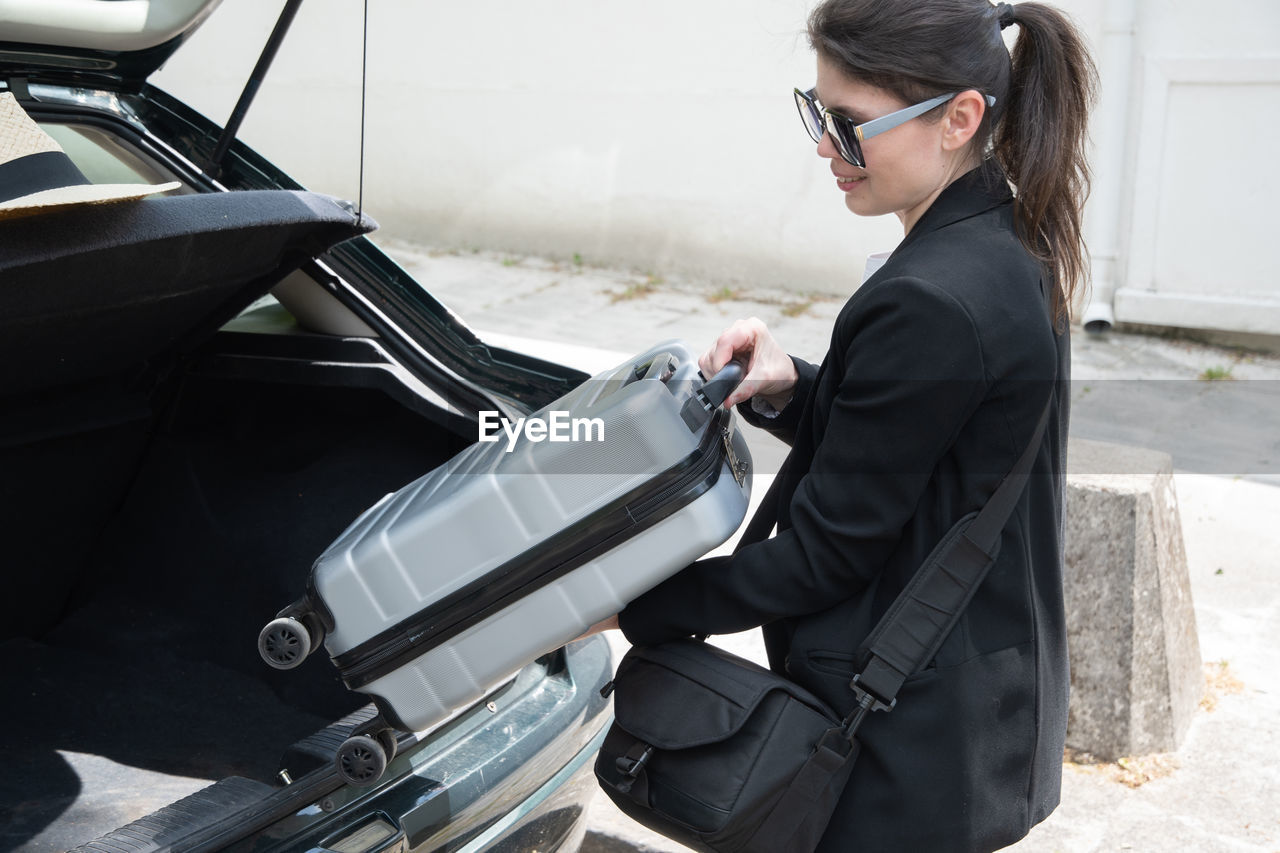 Pretty young brunette putting suitcase in car trunk before going on vacation