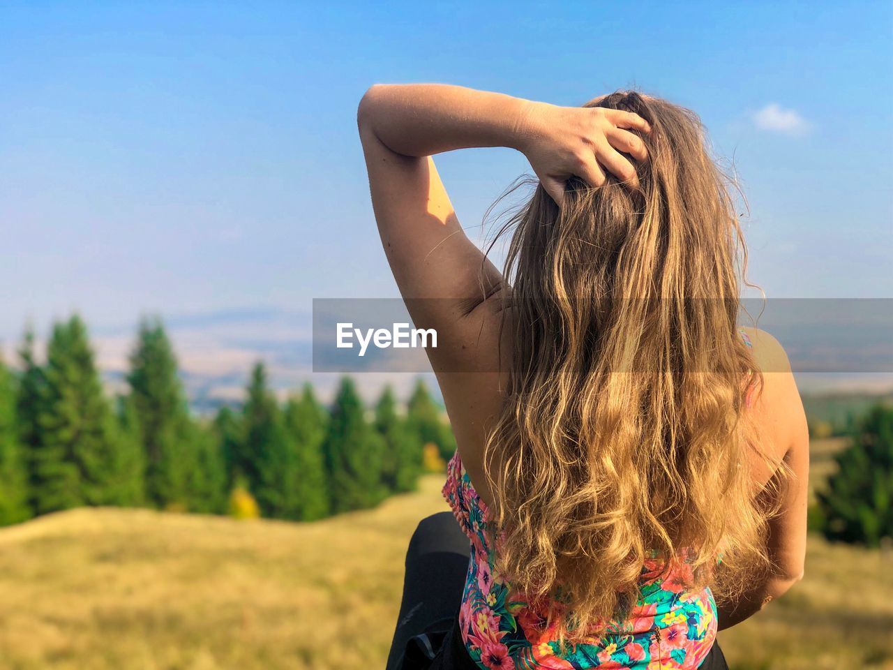 Rear view of mid adult woman with brown hair sitting on grassy field against sky during sunny day