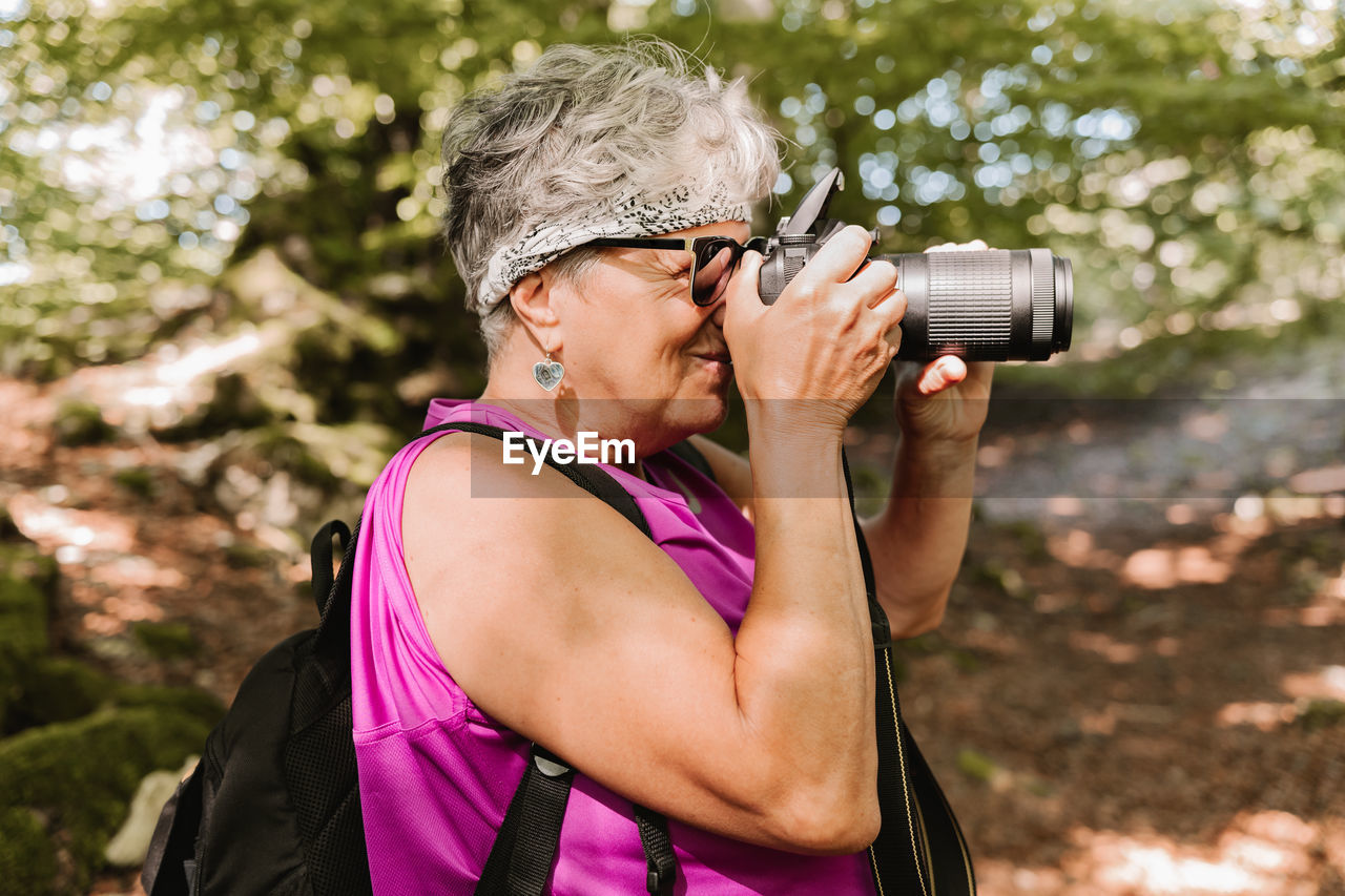 Side view of elderly female tourist with backpack shooting nature while spending sunny summer day in park