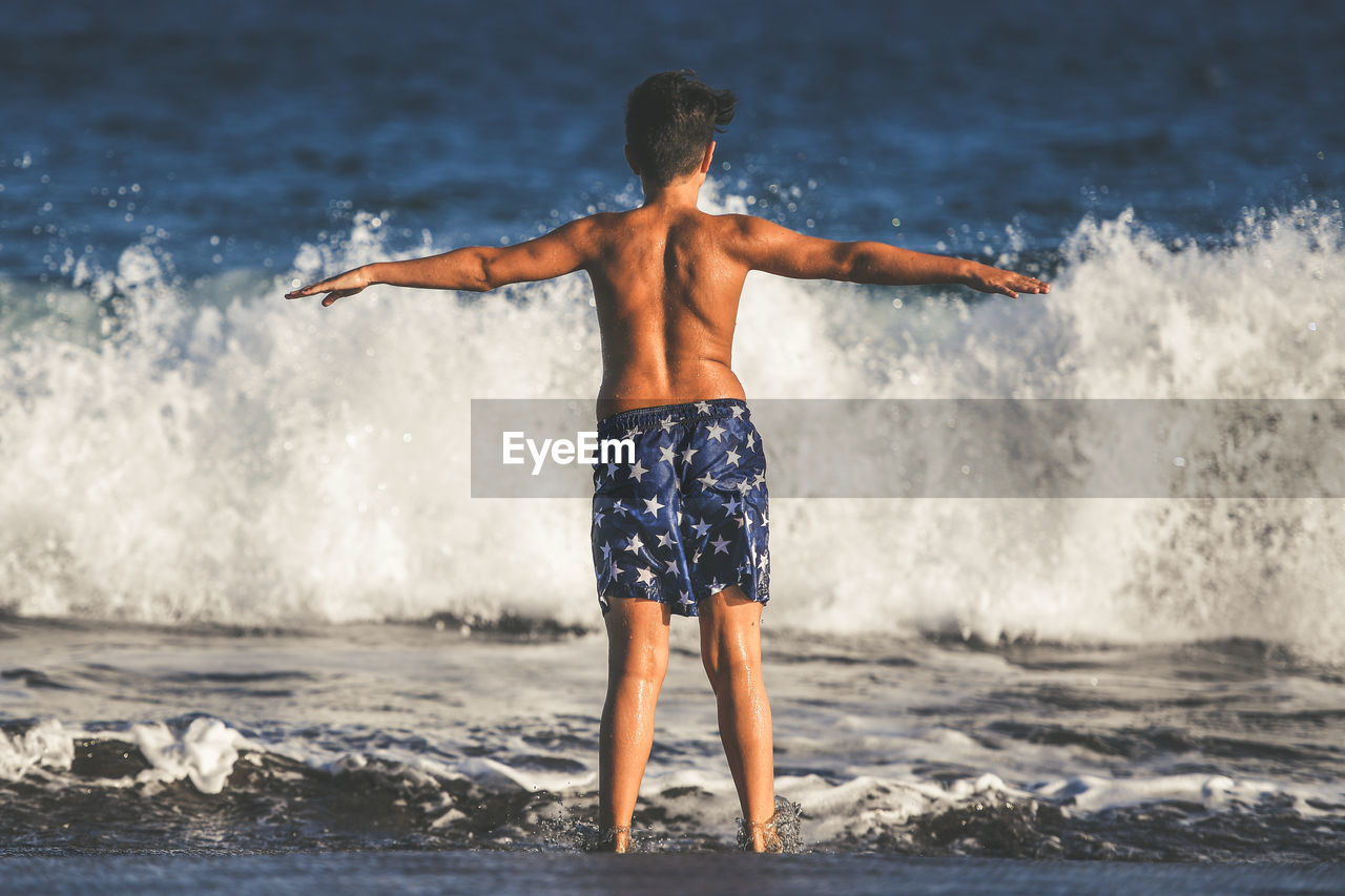 Rear view of shirtless boy with arms outstretched standing on shore at beach