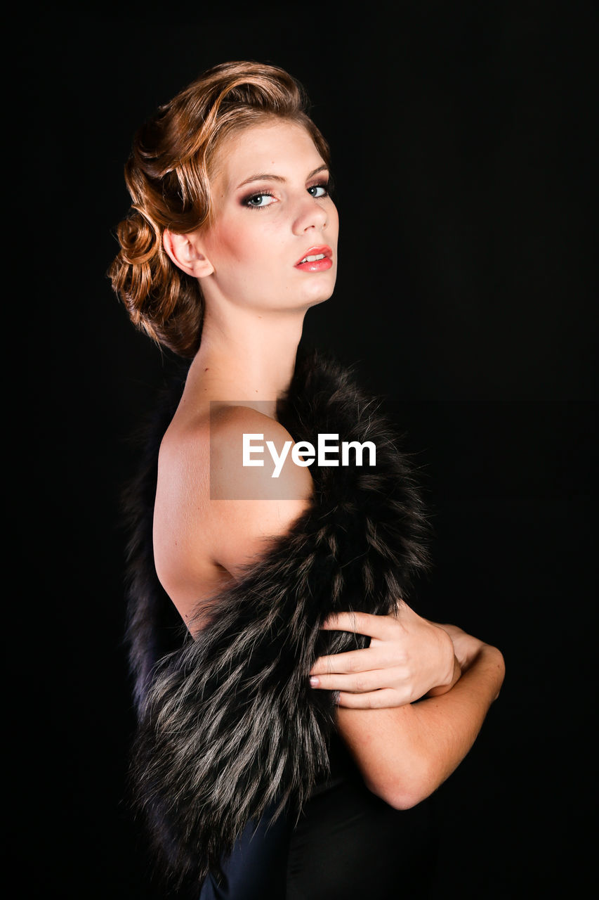 Elegant young woman in black lingerie with fur boa posing at studio on black background