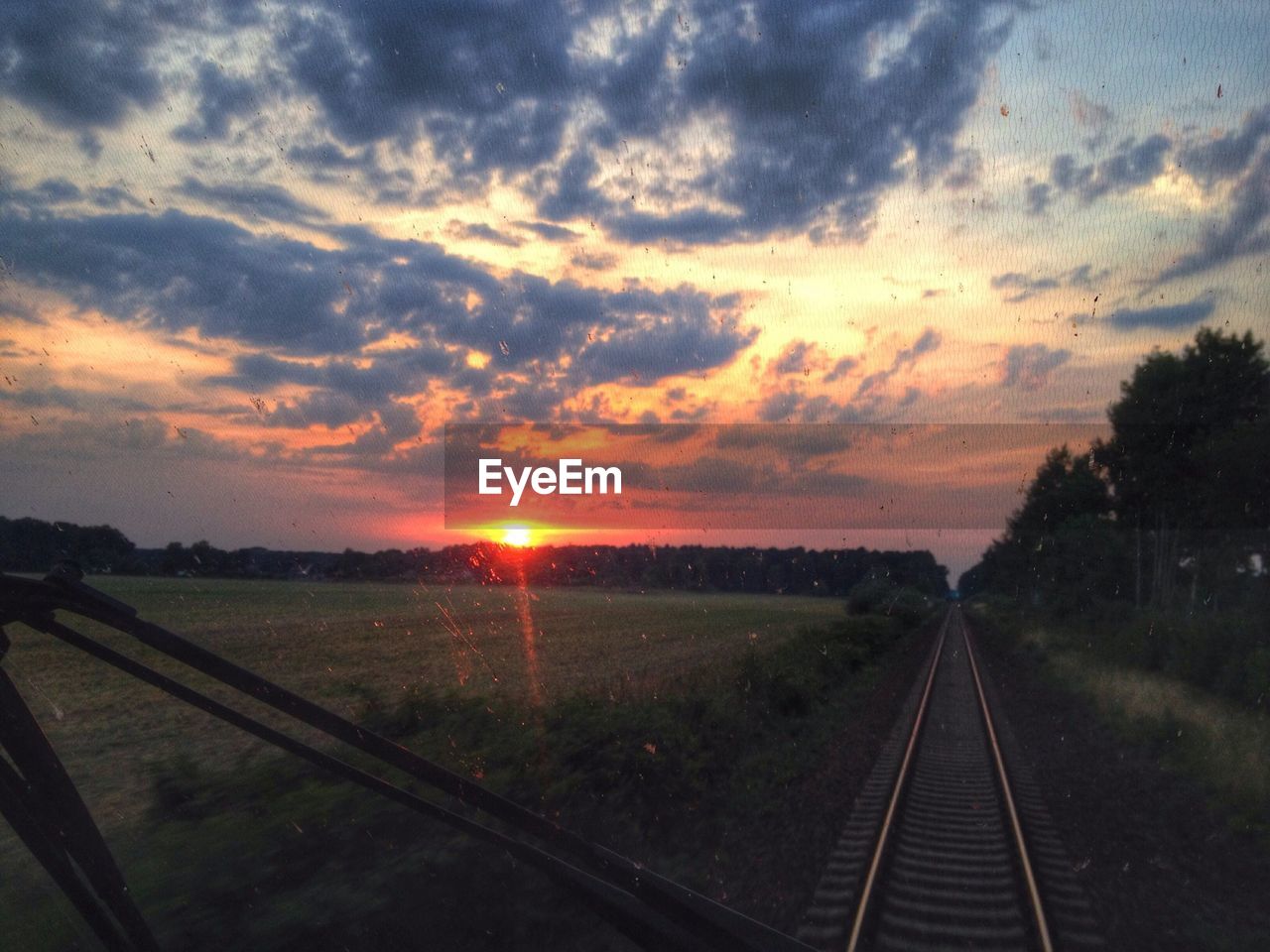 Railroad track amidst field against cloudy sky seen through wet windshield of locomotive