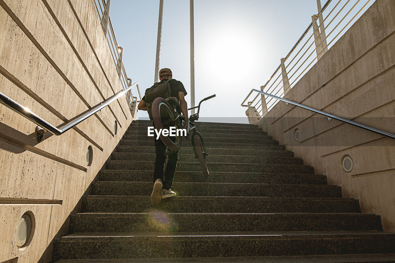 Low angle view of man moving up with bicycle on staircase against sky