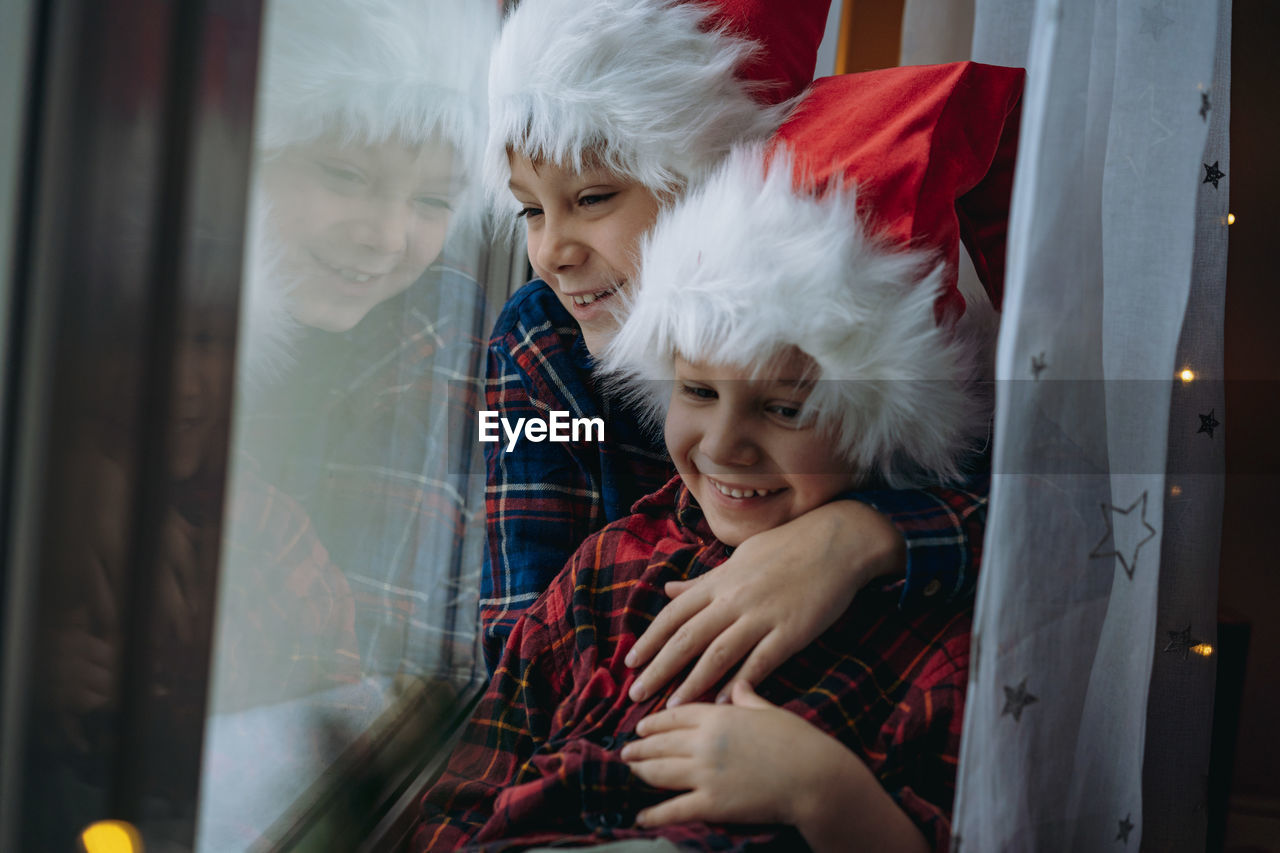 Cute brothers wearing santa hat embracing by window