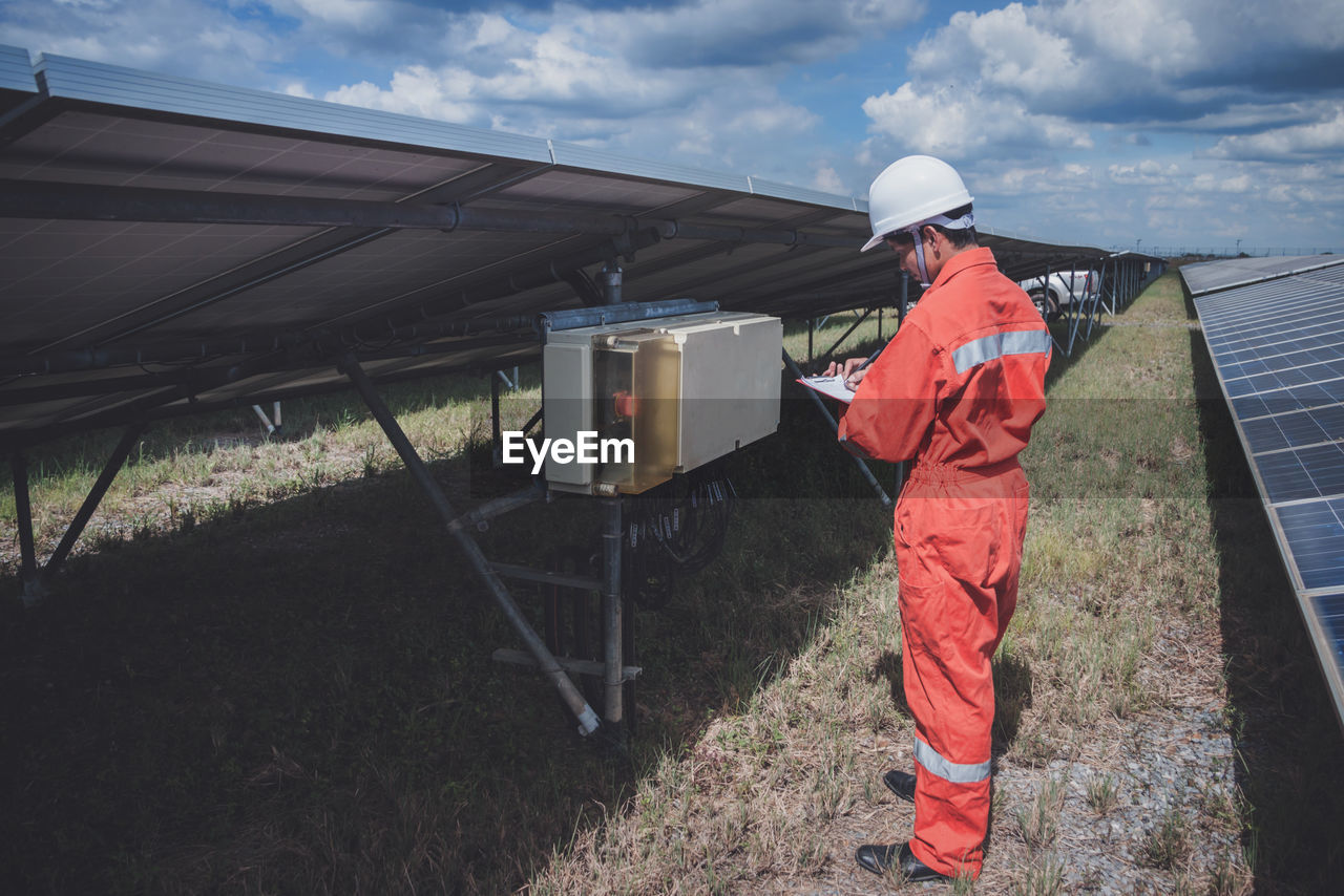 Side view of worker wearing reflective clothing while standing on solar panel against sky