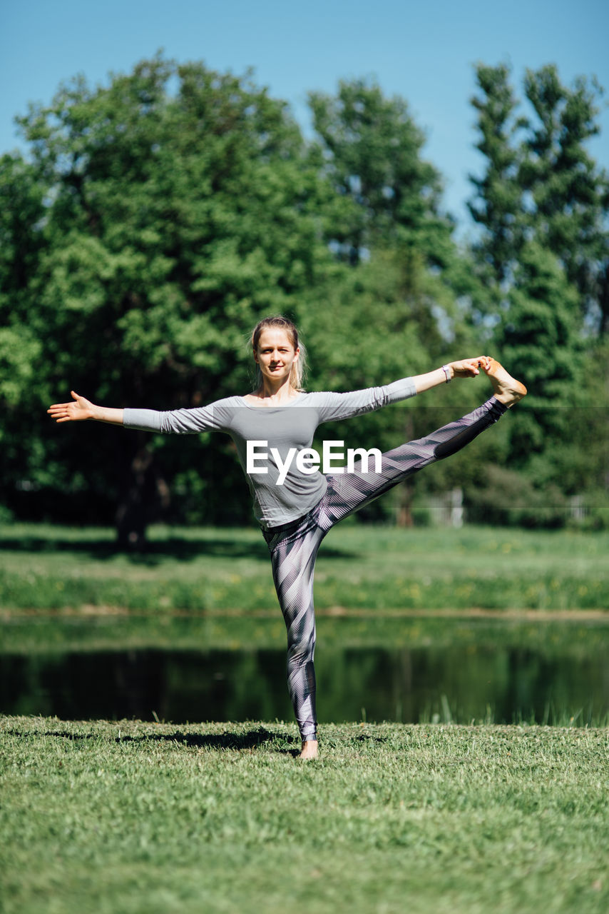 Full length of woman exercising on field by lake at park