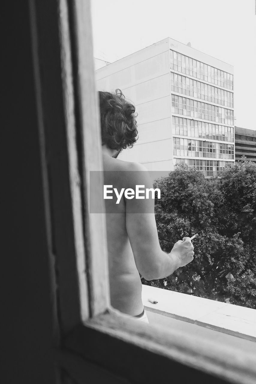 Shirtless man with cigarette standing in balcony seen through window