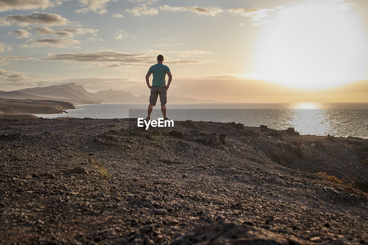 sky, one person, horizon, sea, land, water, beach, sand, ocean, nature, full length, sunlight, beauty in nature, coast, sunset, adult, scenics - nature, standing, cloud, leisure activity, lifestyles, wave, shore, rear view, tranquility, men, vacation, sun, holiday, trip, environment, landscape, solitude, rock, travel, tranquil scene, back lit, idyllic, horizon over water, outdoors, person, exercising, sports, motion, sunbeam, lens flare, relaxation, travel destinations, activity, day, young adult, non-urban scene