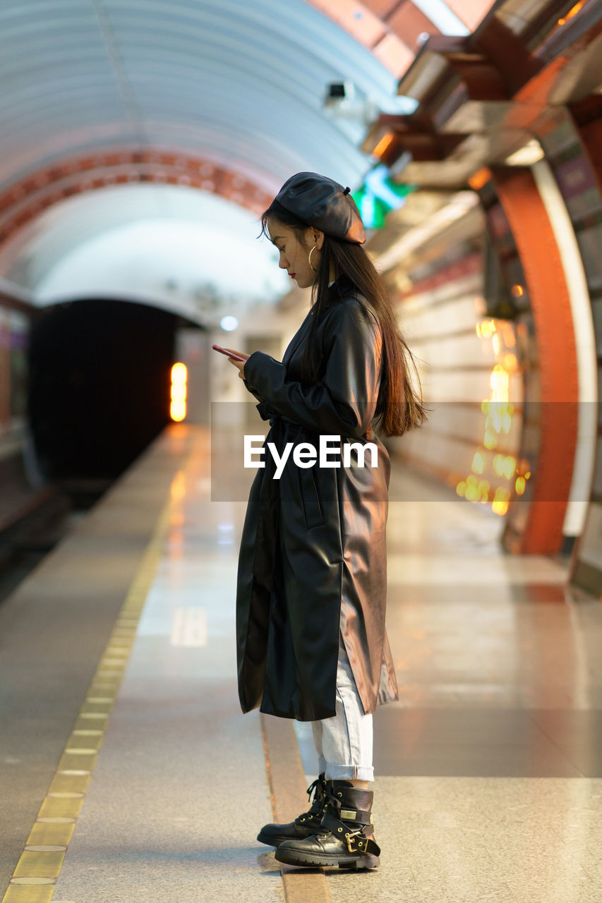 Trendy asian girl wait for train at subway platform check email or online schedule in smartphone