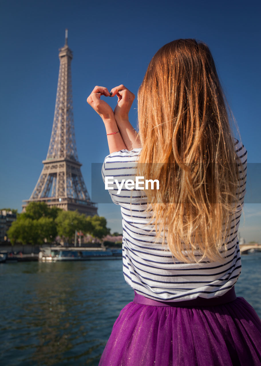 Rear view of woman standing against eiffel tower by river against blue sky