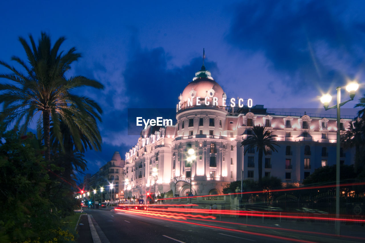 LIGHT TRAILS ON ROAD AMIDST BUILDINGS AGAINST SKY AT NIGHT