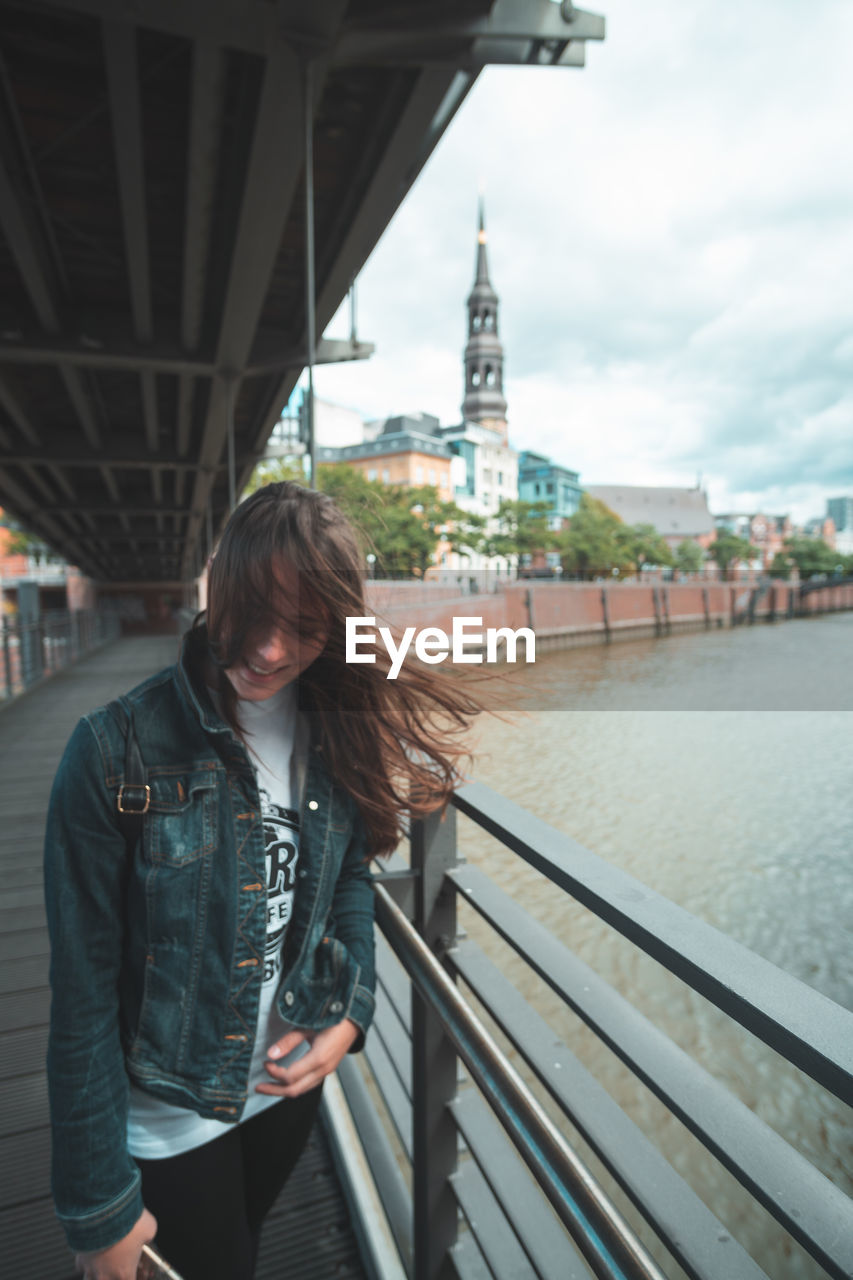 Smiling young woman with tousled hair standing on footbridge by river in city