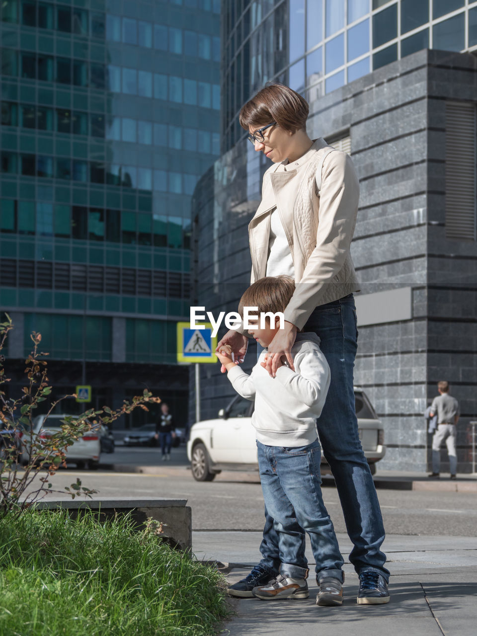 Mother and son stand on street in sunlight. woman and kid in modern town with transport traffic