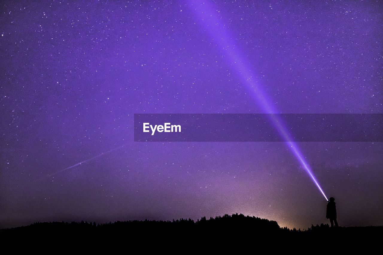 Silhouette person splashing flashlight while standing against purple sky at night