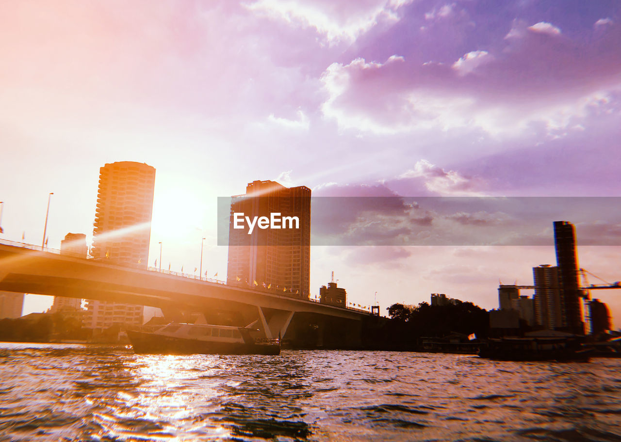 Bridge over river by buildings against sky during sunset