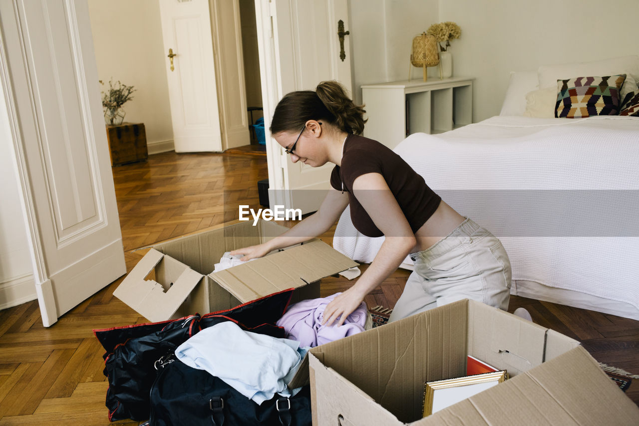 Side view of woman packing clothes in cardboard box while kneeling at home