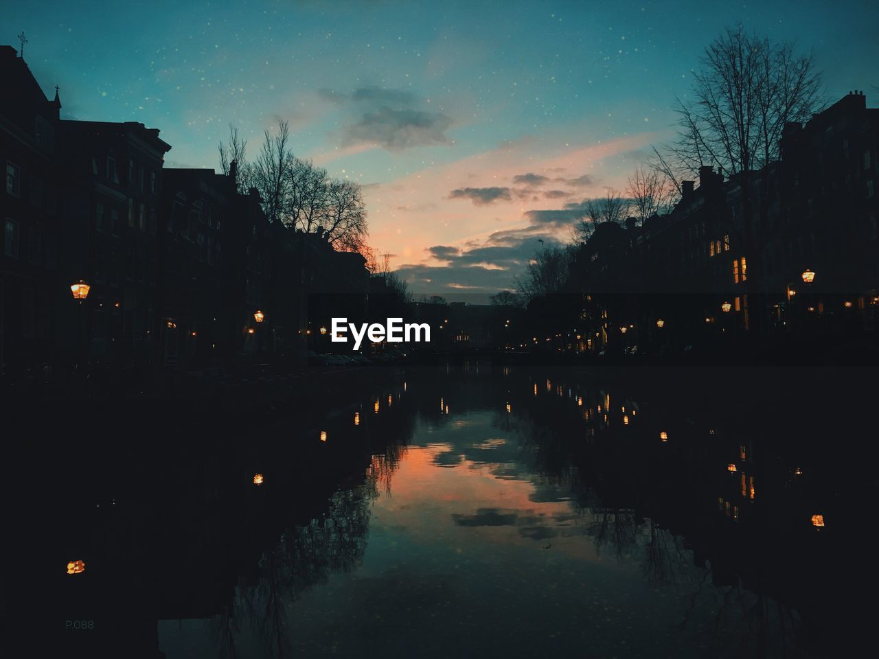 Reflection of sky on calm river amidst houses during sunset