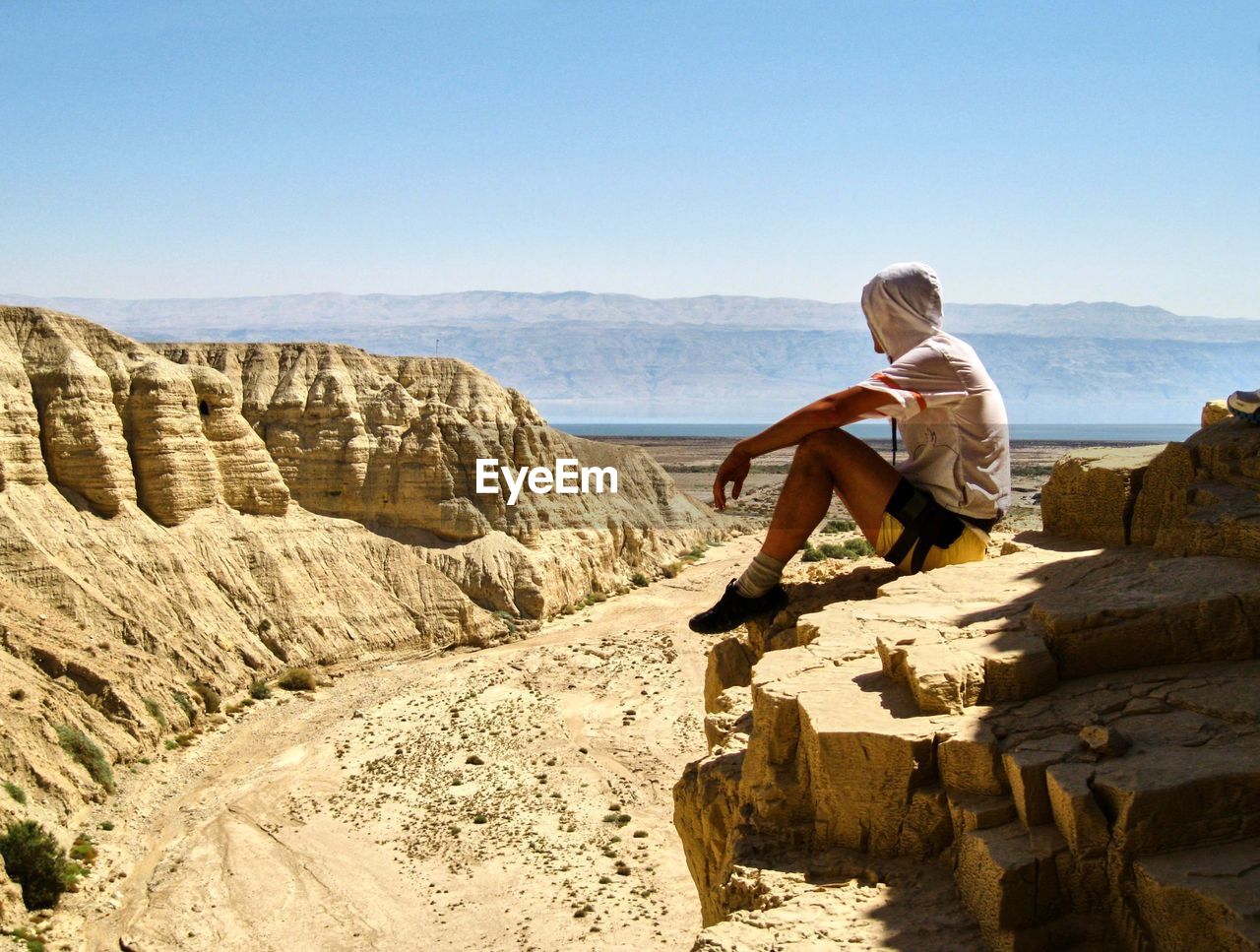 Full length of man sitting on cliff against clear sky during sunny day