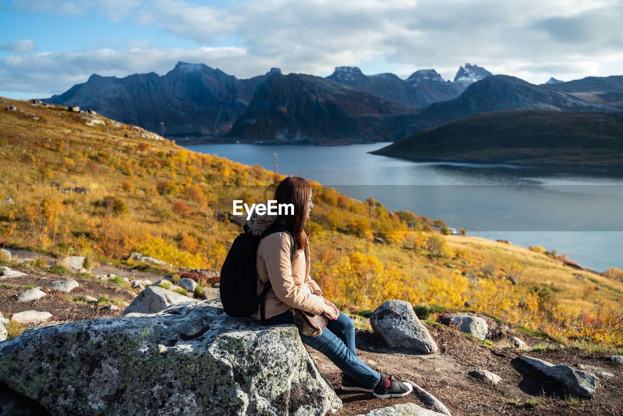 Side view of woman with backpack sitting on rock by lake against cloudy sky