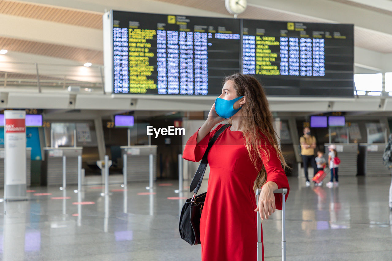 Serious female traveler in mask standing with suitcase on background of departure board and talking on the phone while waiting for flight