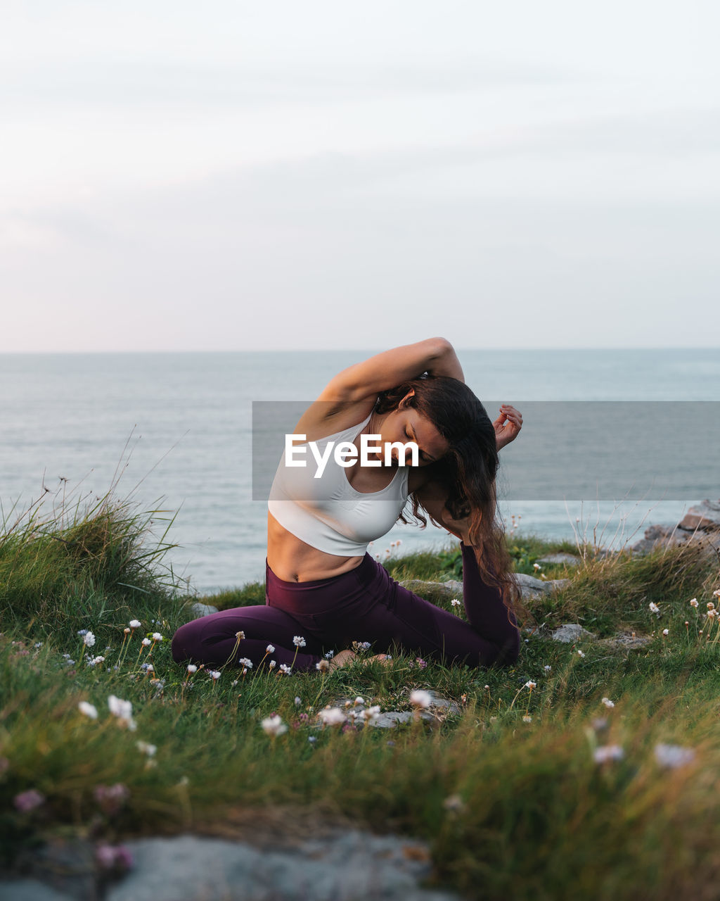 Full length of female athlete in sportswear sitting on grassy hill in eka pada raja kapotasana pose and looking down while practicing yoga in nature