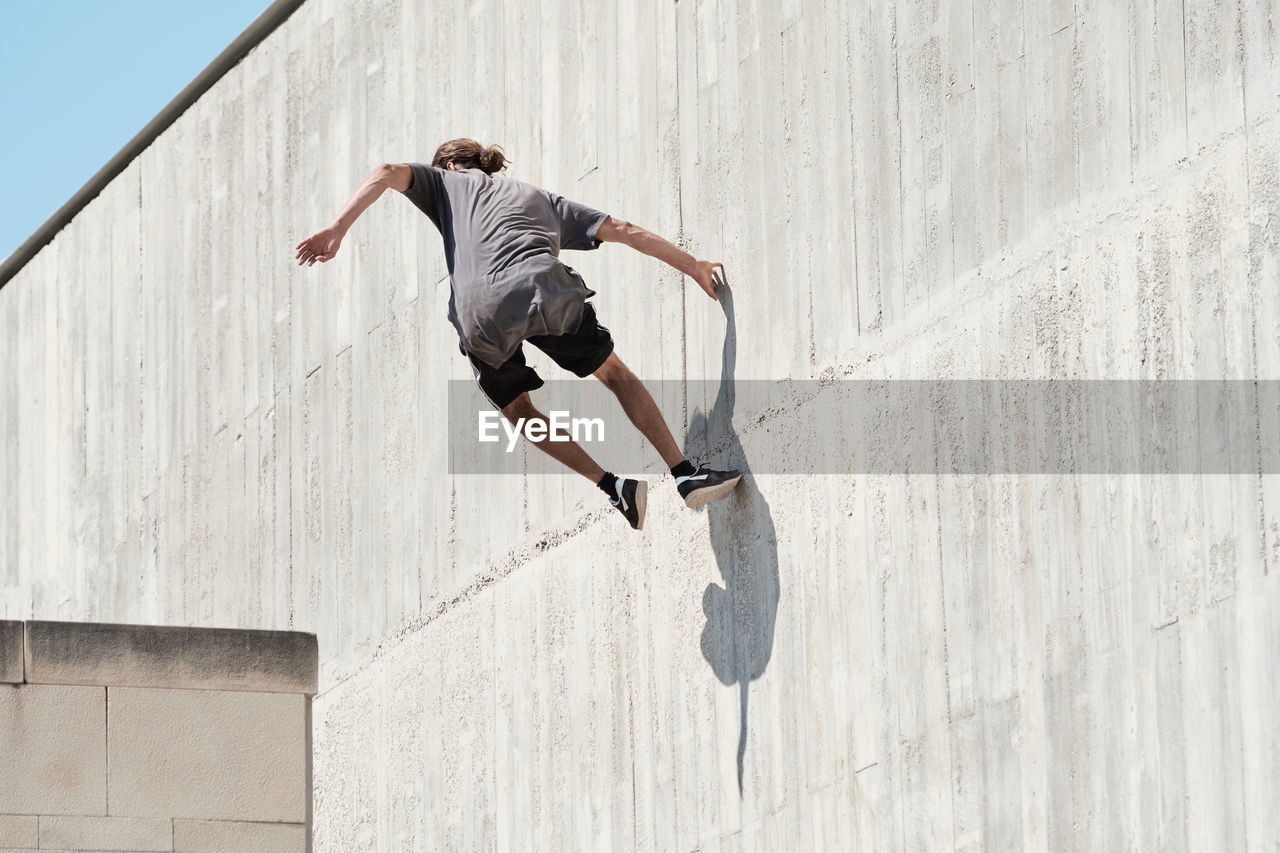Back view of courageous young male jumping on concrete wall of building while performing dangerous stunt and doing parkour in city