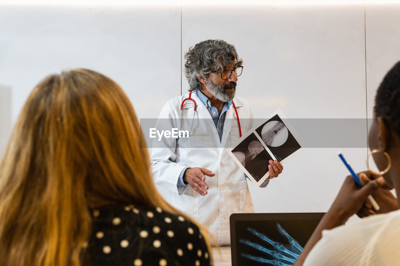 Elderly male medic explaining x ray image to unrecognizable coworkers while working in modern clinic