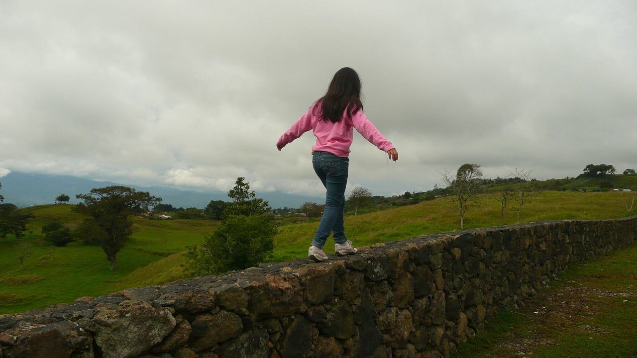 Rear view of woman with arms outstretched walking on retaining wall against cloudy sky