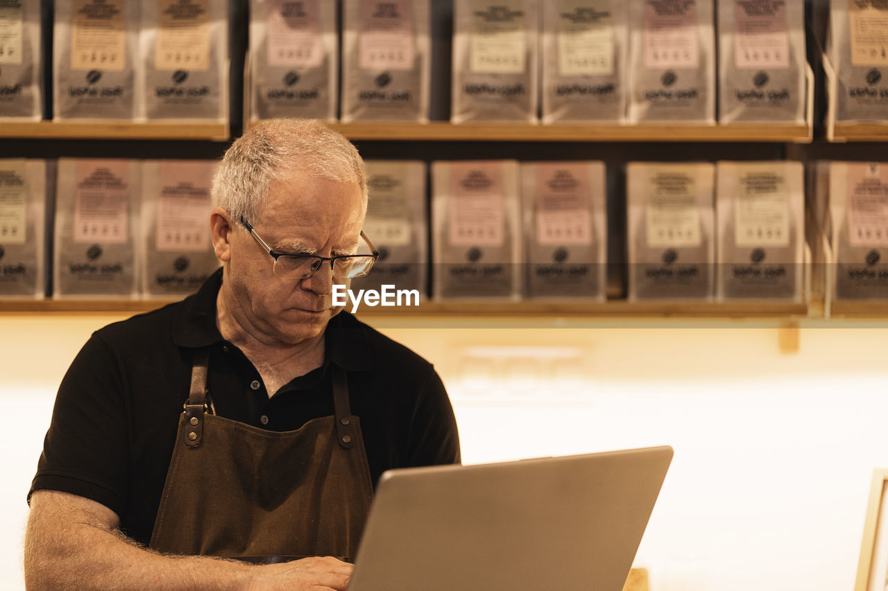 Concentrated mature male barista with gray hair in apron and eyeglasses using laptop while standing at counter in modern coffee shop