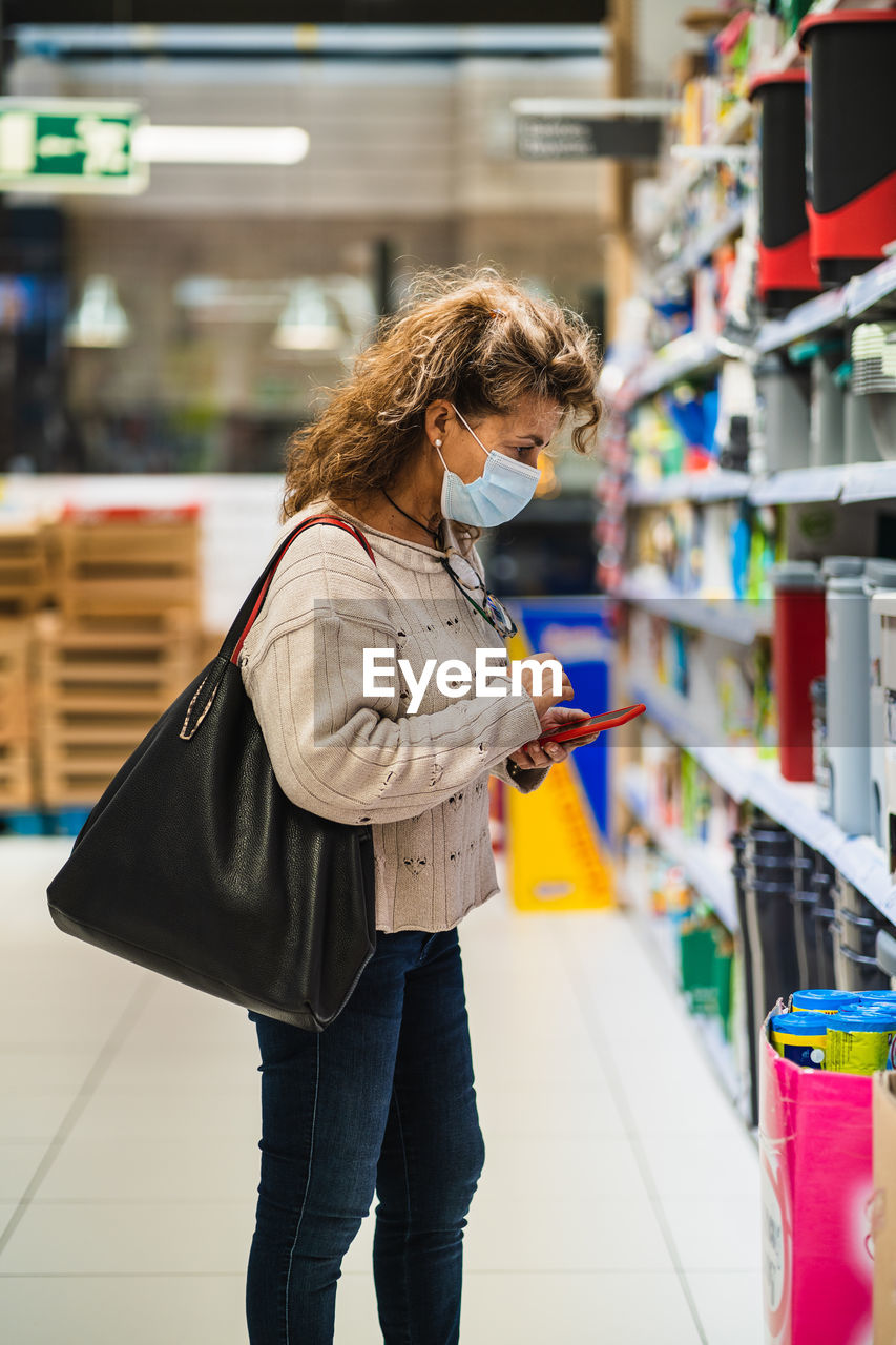 Side view of woman wearing mask using mobile at shopping mall