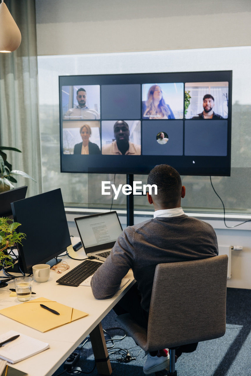 Rear view of businessman doing video conference with colleagues on tv screen in office