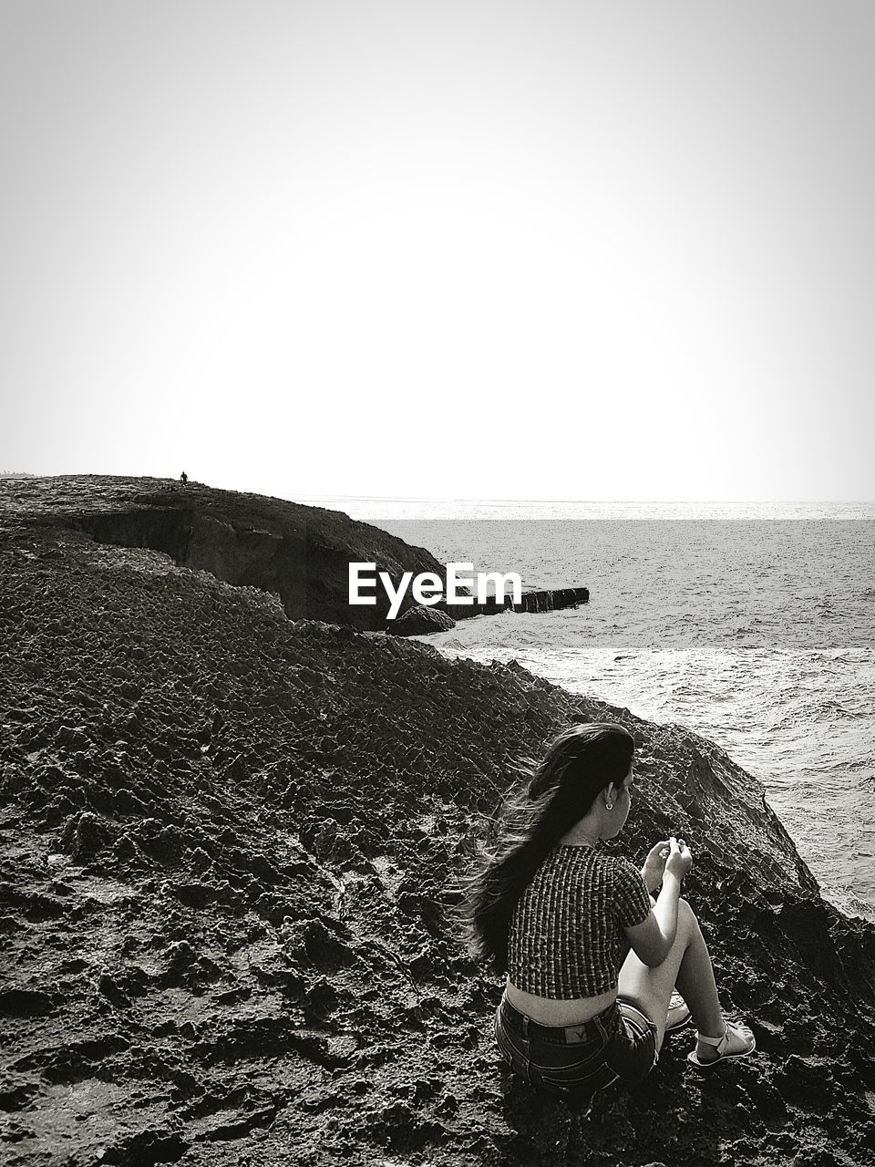 REAR VIEW OF PEOPLE SITTING ON ROCK AGAINST SEA