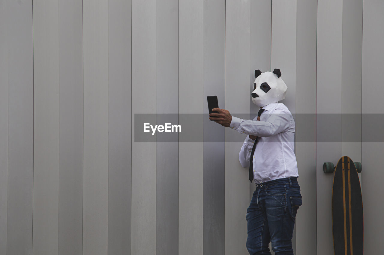 Man in white shirt, tie and panda mask taking smartphone selfie against wall