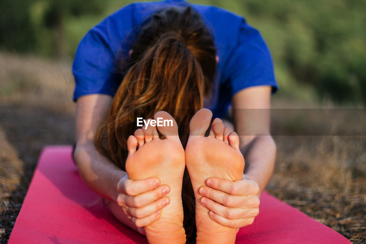 Anonymous flexible female sitting barefoot on mat at sunset and doing yoga in pascimottanasana while practicing mindfulness