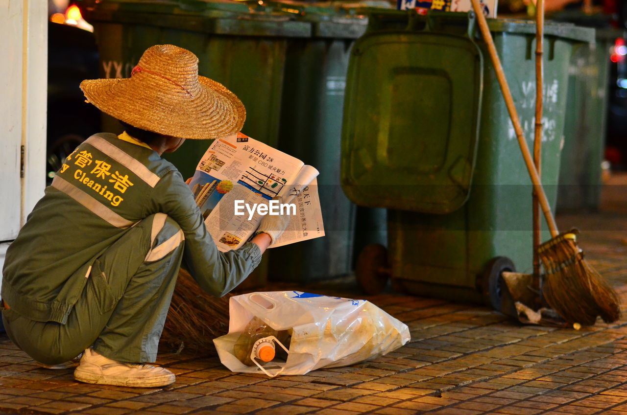 Man wearing straw hat crouching while reading book near dustbin