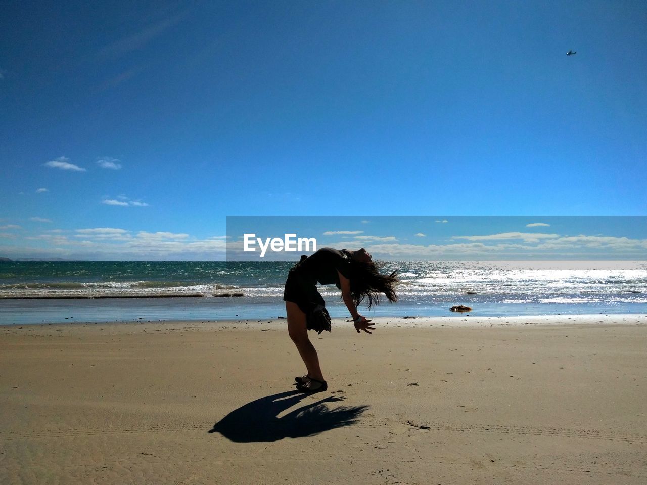 Playful young woman bending over backwards at beach against blue sky