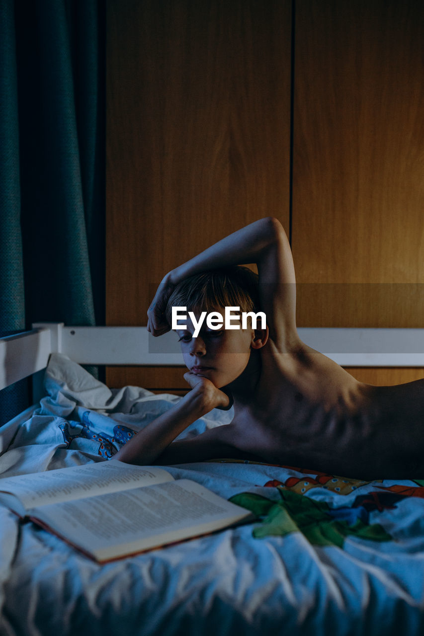 Shirtless boy reading book while lying on bed at home