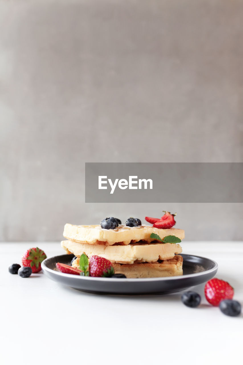 Stack of waffles on a plate on the white table with blueberry, chopped strawberry and mint leaves