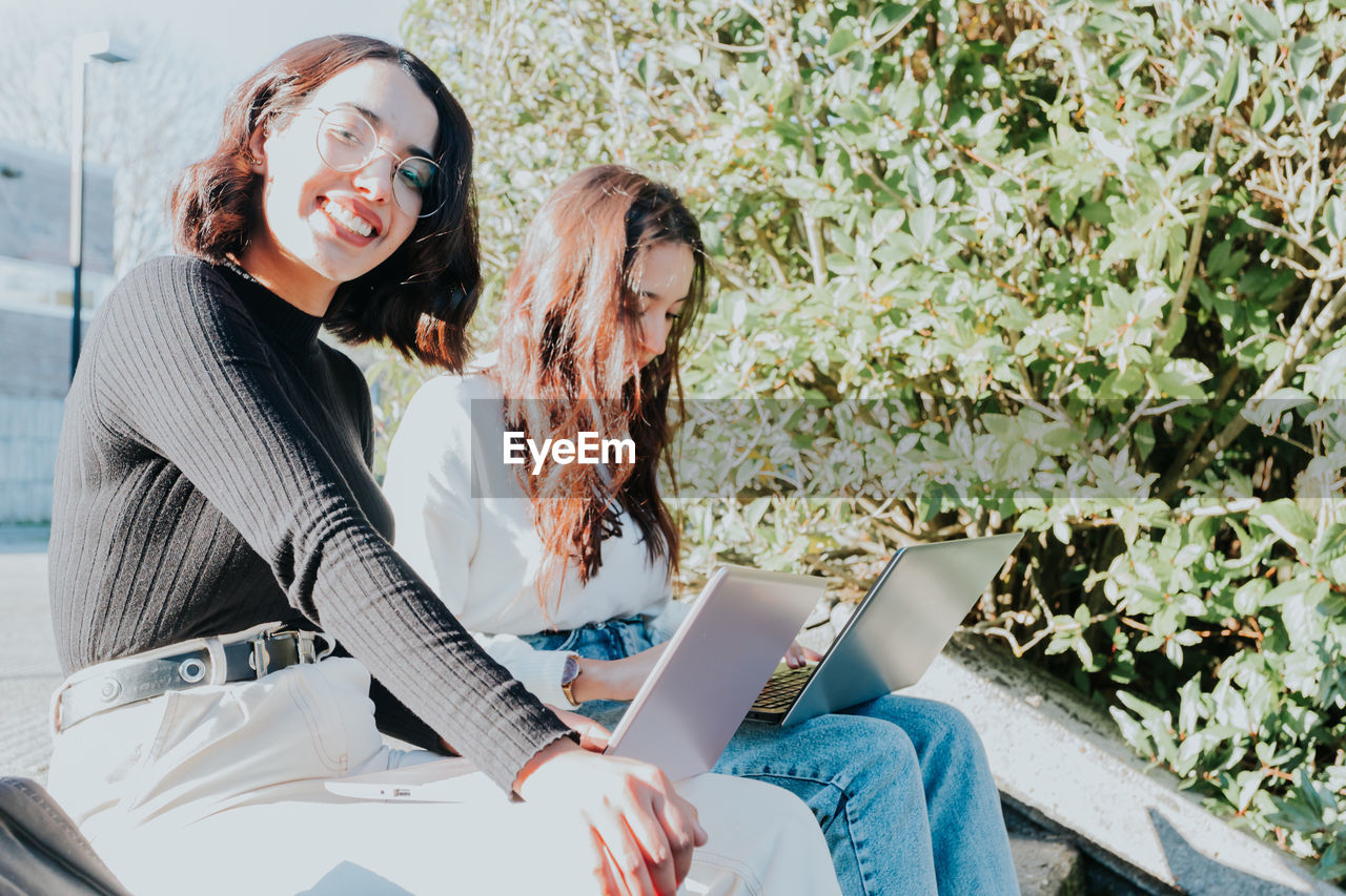 Portrait of smiling businesswoman with laptop by friend on sunny day