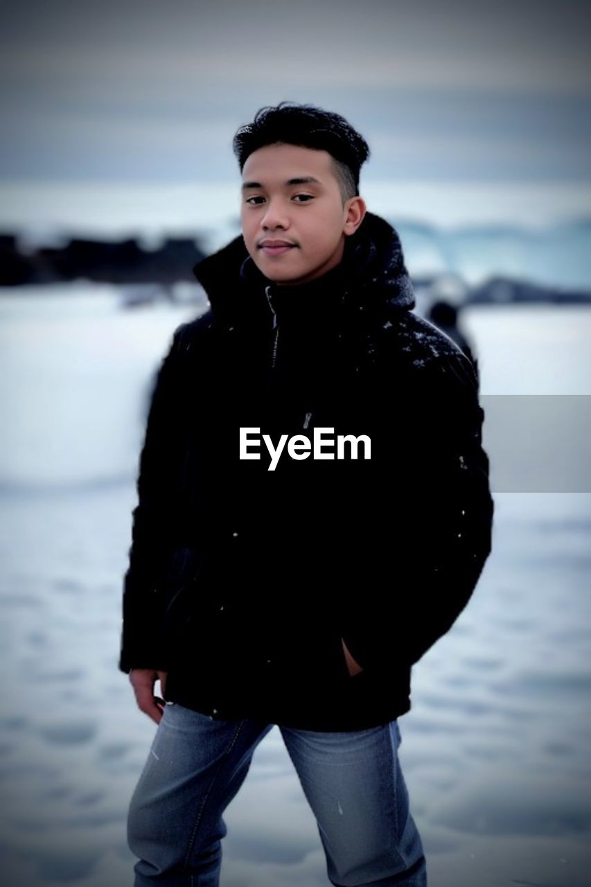 one person, winter, snow, black, cold temperature, clothing, young adult, blue, nature, portrait, water, adult, standing, three quarter length, warm clothing, looking at camera, sky, fashion, photo shoot, front view, focus on foreground, sea, outdoors, person, emotion, leisure activity, looking, land, beauty in nature, portrait photography, smiling, jacket, women, hat, casual clothing