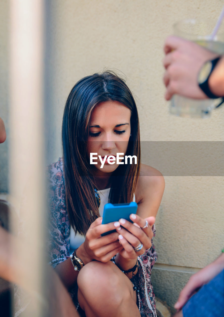 Young woman using mobile phone while sitting against wall