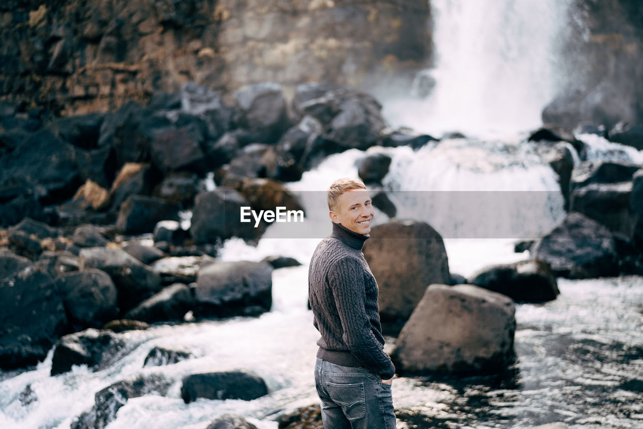 PORTRAIT OF MAN STANDING ON ROCK AGAINST WATERFALL