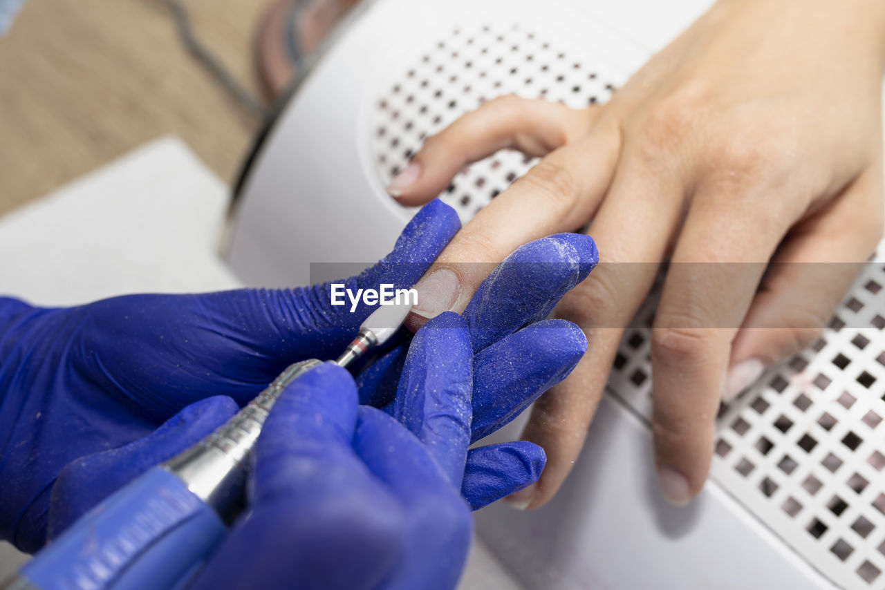 The manicurist grinds the nail plate with a milling machine in blue latex gloves.