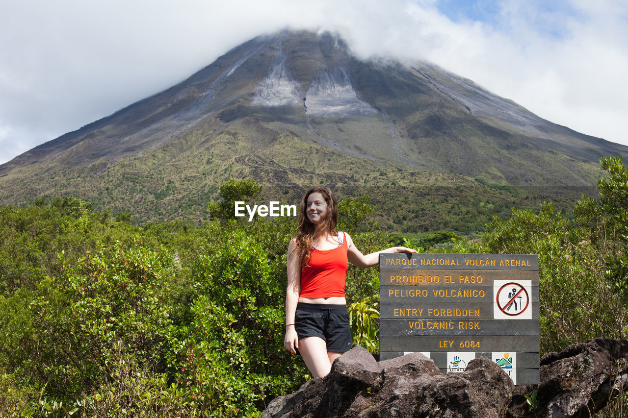 Smiling young woman standing by sign against volcano