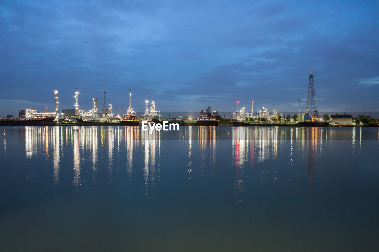 PANORAMIC VIEW OF ILLUMINATED FACTORY BY SEA AGAINST SKY