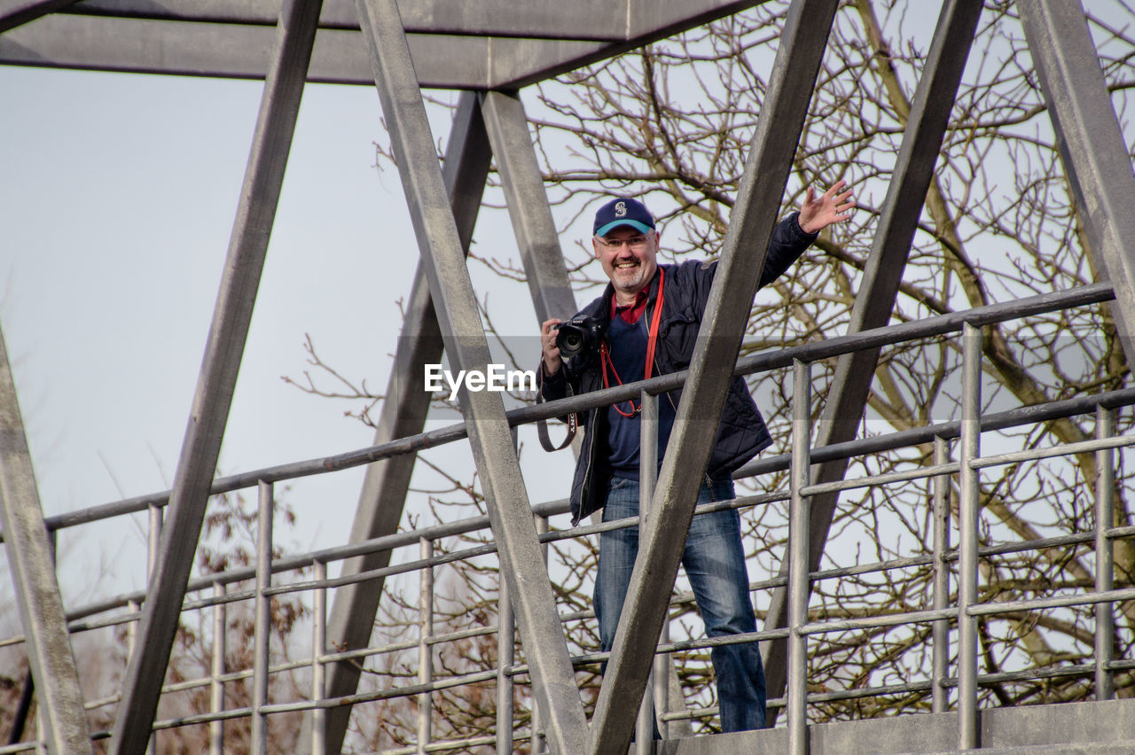 Smiling man with camera gesturing while standing on bridge against bare trees